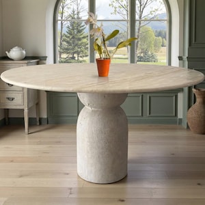 May include: A large, oval-shaped dining table with a light beige marble top and a cylindrical, textured base. An orange pot with a plant sits on the table. The table is in a room with a large arched window.