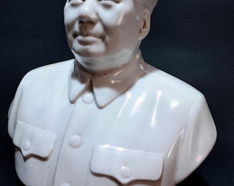 May include: White porcelain bust of a man with short, dark hair. The bust is of a man with a serious expression. The bust is on a black background. The bust has the text "主席万岁!" on the base.