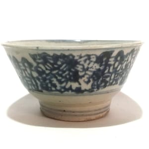 Op de afbeelding: Een witte keramische kom met een blauw bloemmotief rond de rand. De kom heeft een voet en een licht uitlopende vorm. Het blauwe patroon is een herhalend ontwerp van gestileerde bloemen of bladeren. De kom is waarschijnlijk vintage of antiek.