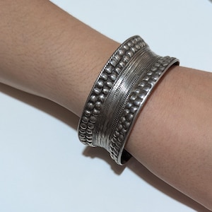 Puede incluir: Un brazalete ancho de plata con detalles intrincados. El brazalete presenta filas de pequeños puntos en relieve y bandas texturizadas. El metal tiene un aspecto ligeramente deslustrado, lo que sugiere un estilo antiguo o vintage. El brazalete se lleva en la muñeca.
