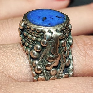 Peut inclure: Une bague en argent avec une grosse pierre ovale bleu foncé. La monture de la bague présente un motif texturé avec de petits ornements ronds et des détails floraux, créant une esthétique vintage. La pierre a des mouchetures sombres.