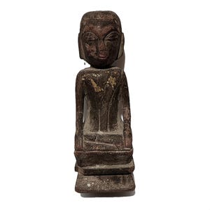 May include: A weathered, wooden statue of a seated figure with a serene expression. The statue is carved from dark brown wood, showing signs of age and wear. The figure is seated with hands resting on knees, and the head is slightly tilted.