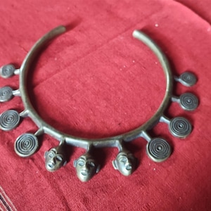 Puede incluir: Collar antiguo de color bronce con un diseño circular. El collar presenta espirales y tres pequeños colgantes con forma de rostro humano. El collar se exhibe sobre una superficie de tela roja.