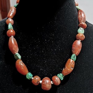 Puede incluir: Un collar con una combinación de cuentas de color marrón rojizo y turquesa. Las cuentas tienen varias formas, como ovaladas y redondas, y están ensartadas para crear una pieza de joyería única. El collar se muestra sobre un fondo negro.