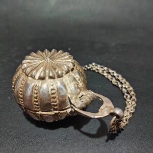 May include: A silver, ornate, round, hinged box with a chain attached. The box has a decorative, floral design.