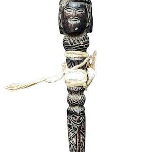 May include: A carved wooden figure with a human face and intricate details. The figure is tied with a white string and has a dark brown color.