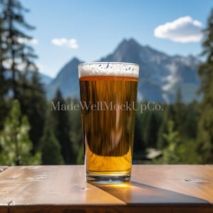 May include: A glass of beer with a white head sits on a wooden table with a mountain range in the background.