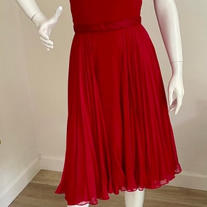 May include: A red sleeveless dress with a sheer overlay at the neckline and a black ribbon belt. The dress has a fitted bodice and a flowing skirt.