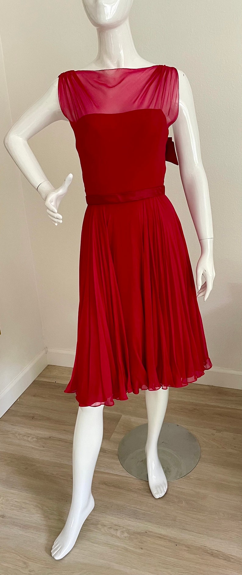 May include: A red sleeveless dress with a sheer overlay at the neckline and a black ribbon belt. The dress has a fitted bodice and a flowing skirt.
