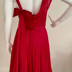 May include: A fuchsia pink pleated dress with a large bow at the back. The dress has a scoop neckline and a fitted bodice. The skirt is full and flowing.