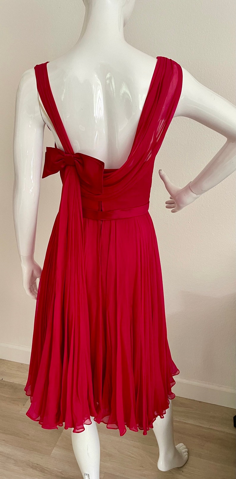 May include: A fuchsia pink pleated dress with a large bow at the back. The dress has a scoop neckline and a fitted bodice. The skirt is full and flowing.