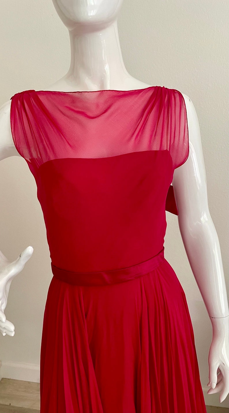 May include: A sleeveless, fuchsia-coloured dress with a sheer, ruffled neckline and a fitted bodice. The dress has a wide, satin belt and a flowing skirt.