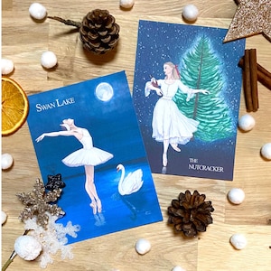 Puede incluir: Dos tarjetas ilustradas con bailarinas. La primera tarjeta muestra una bailarina con un tutú blanco bailando con un cisne en un cielo azul y estrellado. La tarjeta dice "Swan Lake". La segunda tarjeta muestra una bailarina con un vestido blanco bailando frente a un árbol de Navidad verde. La tarjeta dice "El Cascanueces".