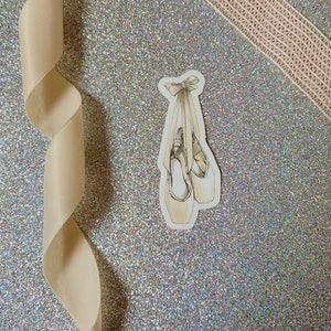 Peut inclure: Un autocollant de chaussure de ballet blanche avec un nœud sur un fond de paillettes argentées.