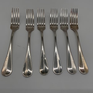 Könnte beinhalten: Sechs silberne Gabeln mit klassischem Design sind auf einer hellgrauen Oberfläche angeordnet. Jede Gabel hat einen langen, schlanken Griff und vier Zinken. Die Gabeln sind gleichmäßig verteilt und reflektieren das Licht.