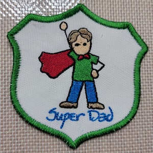 May include: Green and white embroidered patch with a cartoon image of a person wearing a red cape and blue pants. The text "Super Dad" is embroidered in blue below the image.