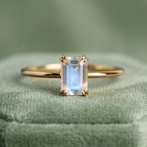 Puede incluir: Un anillo de oro con una piedra preciosa rectangular e iridiscente. La piedra preciosa tiene un color azul y blanco. El anillo está sobre una superficie de terciopelo verde.