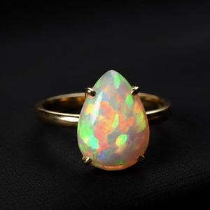 May include: A gold ring featuring a pear-shaped opal gemstone. The opal displays a vibrant play of color with hues of green, orange, and yellow. The ring is set against a black background, highlighting the gemstone's brilliance.