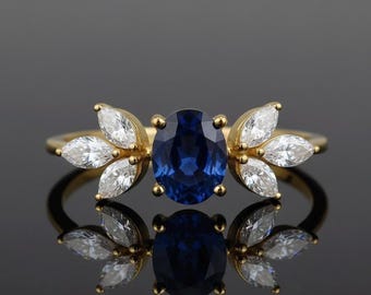 May include: A gold ring featuring a large, oval, deep blue sapphire center stone. Flanking the sapphire are six marquise-cut, white gemstones, arranged in a leaf-like design. The ring is set against a dark, reflective surface.