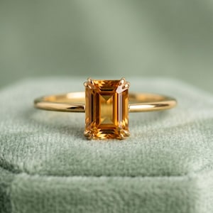 May include: A gold ring with a rectangular, amber-colored gemstone. The ring's band is thin and polished, and the gemstone is set with four prongs. The ring is displayed on a soft, green surface.