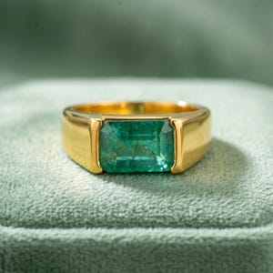 May include: A gold ring with a rectangular emerald gemstone. The emerald is a vibrant green color and is set in a gold band. The ring is displayed on a green velvet surface.