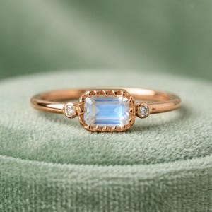 Könnte beinhalten: Ein roségoldener Ring mit einem rechteckigen, hellblauen Edelstein in der Mitte. Der Edelstein ist von einer dekorativen Bordüre umgeben. Kleine, runde, klare Edelsteine ​​sind auf beiden Seiten des Hauptsteins gesetzt.