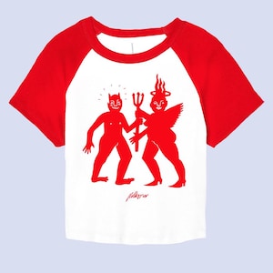 May include: White cropped t-shirt with red raglan sleeves. The shirt features a red illustration of two figures with horns and wings, one holding a trident. The text "Pillows" is printed below the illustration.
