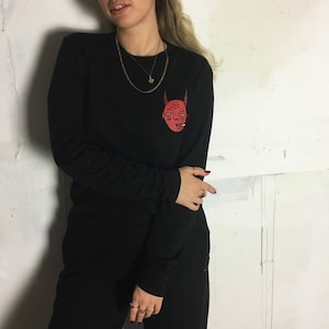 May include: Black long-sleeve shirt with a red cartoon face graphic on the front. The face has horns and a cigarette in its mouth.