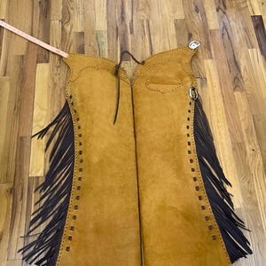 May include: A pair of brown leather chaps with fringe and stitching detail. The chaps have a buckle and strap on the side.