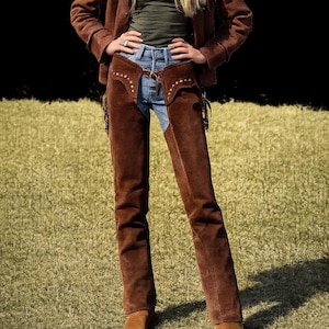 May include: A person wearing a brown suede jacket, a green top, blue jeans, and brown suede chaps with decorative studs. The person is also wearing brown suede boots. The background is a grassy field.