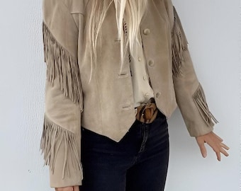 May include: A beige suede jacket featuring long fringe detailing along the sleeves, a collared neckline, and a front button closure. The jacket is paired with a light-colored button-down shirt, a brown belt with a circular buckle, and black pants.