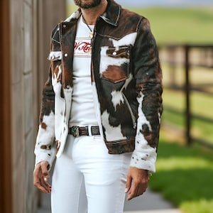 May include: A cowhide jacket with brown, white, and black patterns. The jacket has a classic collar, buttoned cuffs, and two chest pockets. The person is wearing a white t-shirt with text and white trousers. A brown belt with a decorative buckle is visible.
