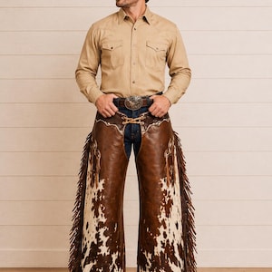 Op de afbeelding: Een man draagt een bruine cowboyhoed, een beige overhemd met knopen en chaps van bruin en wit koeienhuid met franjes. De chaps worden over een blauwe spijkerbroek gedragen. Een zilveren gesp is zichtbaar.