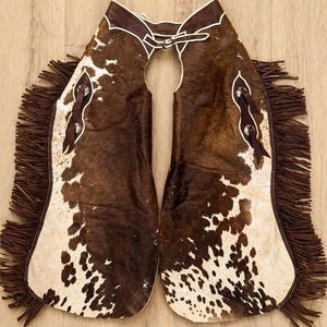 May include: Cowhide chaps with a brown and white spotted pattern. The chaps have brown leather trim, fringe along the sides, and silver-toned hardware. These western-style chaps are designed to protect the legs.