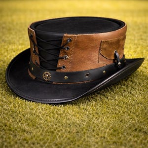 Peut inclure: Un chapeau haut de forme noir avec des détails en cuir marron et un design steampunk. Le chapeau présente un ornement en forme de roue dentée et un laçage latéral. Il repose sur de l'herbe.