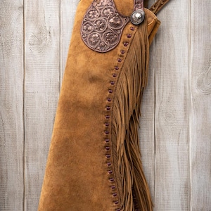 May include: A brown suede leather chaps with decorative floral embossed accents. The chaps feature a fringe along the side and decorative stitching. A silver buckle and concho add detail to the western wear.
