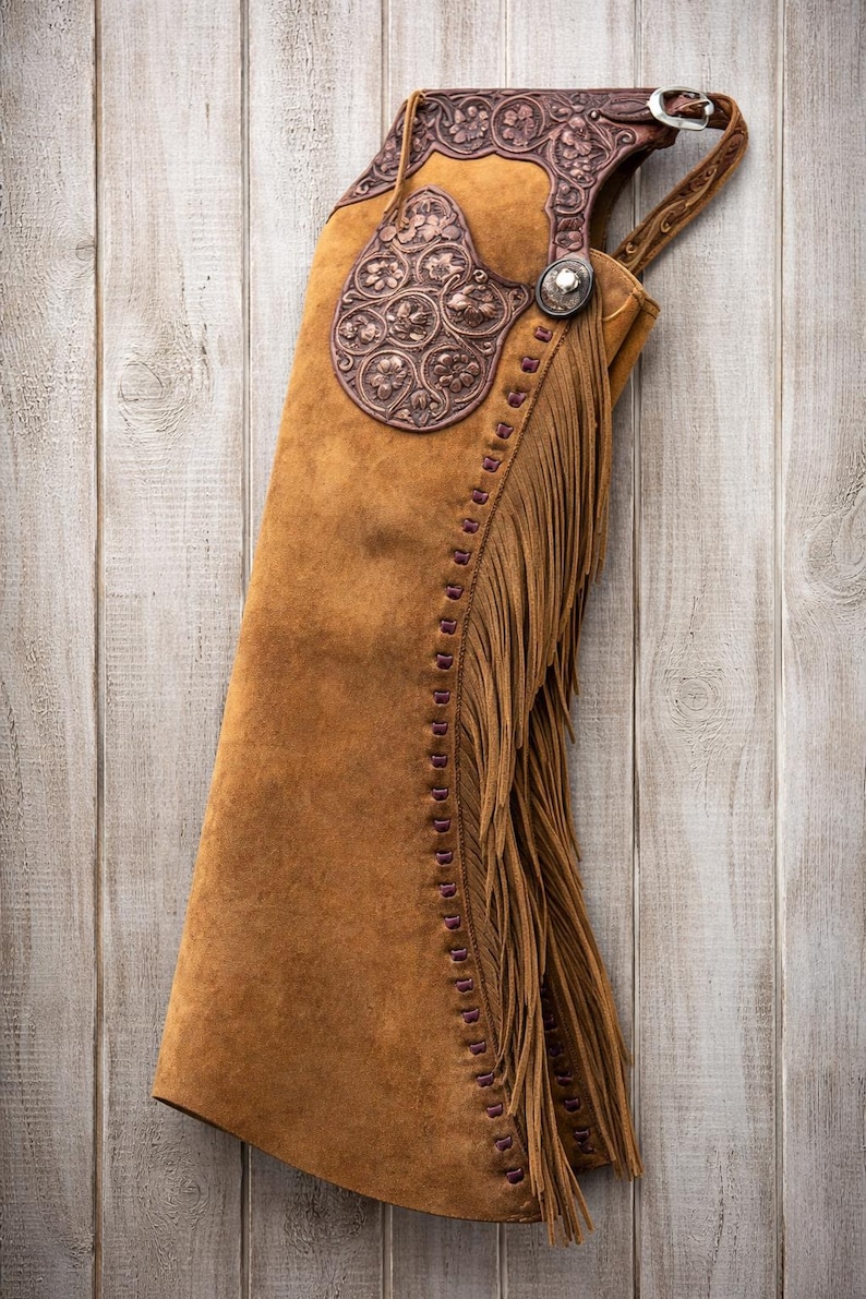 May include: A brown suede leather chaps with decorative floral embossed accents. The chaps feature a fringe along the side and decorative stitching. A silver buckle and concho add detail to the western wear.