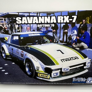 May include: A model of a white, black, and green Mazda Savanna RX-7 Daytona '79 race car. The car has the number 7 on the hood and sponsor logos. The box features the car on a racetrack with people in the background.