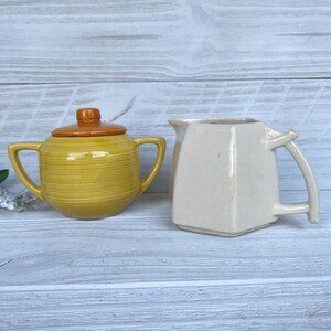 May include: A yellow ceramic sugar bowl with a brown lid and two handles, next to a cream-colored pitcher with a handle. The sugar bowl has horizontal ridges. The pitcher has a square shape.