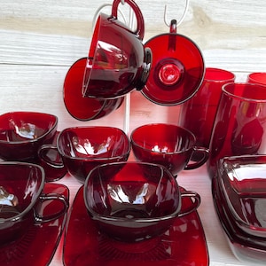 Könnte beinhalten: Eine Sammlung rubinroter Glaswaren, darunter Tassen, Untertassen und Becher. Die Tassen haben quadratische Schalen und abgerundete Griffe. Einige Tassen hängen an einem Metallgestell. Die Glaswaren sind auf einer weißen Oberfläche angeordnet.