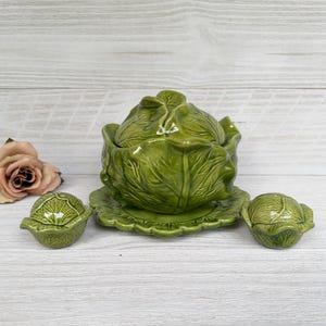 May include: A green ceramic cabbage-shaped serving dish with a lid, a matching saucer, and two smaller cabbage-shaped containers. The set is a vibrant green color and sits on a light-colored wooden surface.