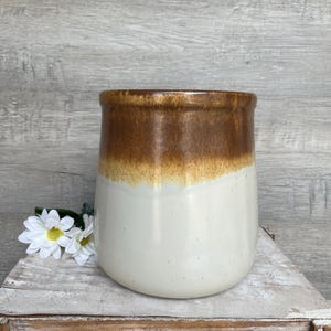 May include: A white ceramic vase with a brown glaze on the top half. The vase has a rounded bottom and a slightly flared top.
