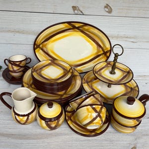 Puede incluir: Un juego de vajilla de cerámica vintage con una base crema y un patrón geométrico amarillo y marrón. El juego incluye platos, tazas, platillos, una fuente para servir, una lechera, una azucarera y una bandeja para servir de dos niveles.