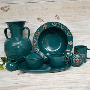 May include: A teal ceramic dinnerware set featuring a large bowl, a serving platter, a creamer, a sugar bowl, a mug, and a decorative vase. The bowl and mug have a brown geometric design. The set is on a light wood surface.