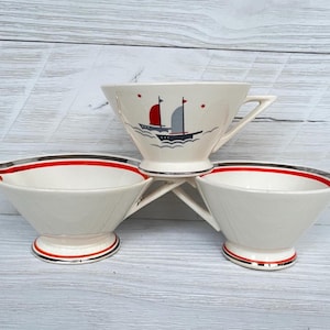 May include: A set of three vintage white ceramic teacups and saucers. The teacup features a sailboat design with red and gray sails. The saucers have red and black stripes around the rim. The set is displayed on a light wood surface.