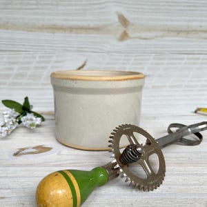 May include: A vintage hand-crank egg beater with a green and yellow wooden handle and a metal gear. A small, off-white ceramic crock with a tan rim sits behind the beater. The items are on a light-colored wooden surface.