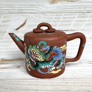 May include: A small, brown ceramic teapot with a dragon design. The dragon is depicted in green, blue, and yellow. The teapot has a curved handle and spout, and a small lid with a loop handle. The teapot is likely vintage or antique.