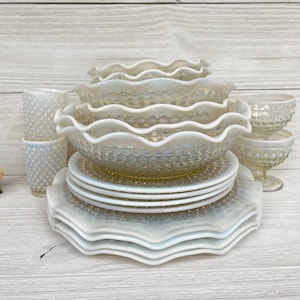 Könnte beinhalten: Eine Sammlung von Vintage-Tafelservice aus opaleszierendem Glas. Das Set umfasst Teller, Schalen und Dessertschalen mit Hobnail-Muster und gewellten Kanten. Das Glasgeschirr ist auf einer hellen Holzoberfläche angeordnet.