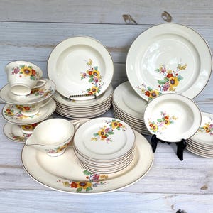 Puede incluir: Un juego de vajilla vintage de color crema con un diseño floral de flores amarillas, rojas y moradas. El juego incluye platos, platillos, tazas, una fuente para servir y una salsera. Cada pieza tiene un borde dorado.