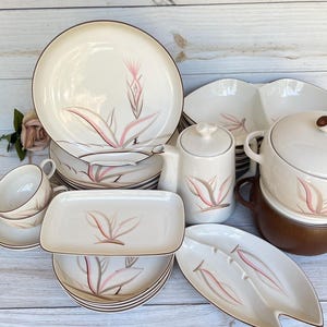 May include: A vintage cream-colored dinnerware set with a floral design in pink, gray, and brown. The set includes plates, bowls, a serving dish, a creamer, a sugar bowl, and a covered casserole dish. The rims are brown.